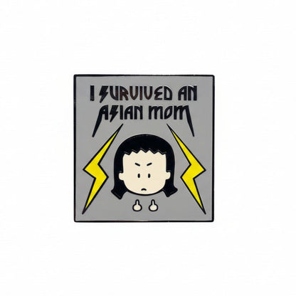 A pin with cartoon character Kim, the Angry Little Asian Girl, and the text 'I SURVIVED AN ASIAN MOM' surrounded by yellow lightning bolts. The pin is attached to a blue surface, and there is a logo and text indicating 'Angry Little Girls by Lela Lee' and 'Anger is a gift' at the bottom.